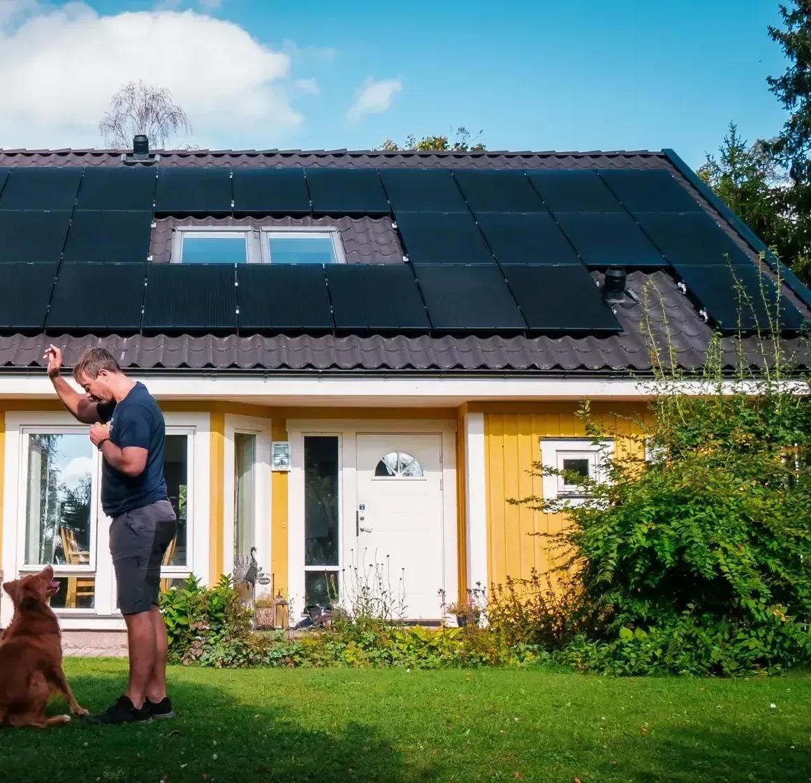 En man och hund framför ett gult hus med svarta solpaneler på taket